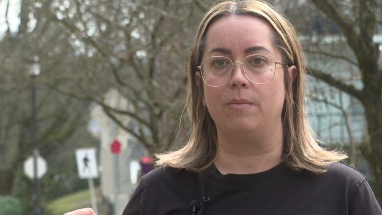 A woman with short blonde hair, round glasses and a black t-shirt stands in a residential neighbourhood. 