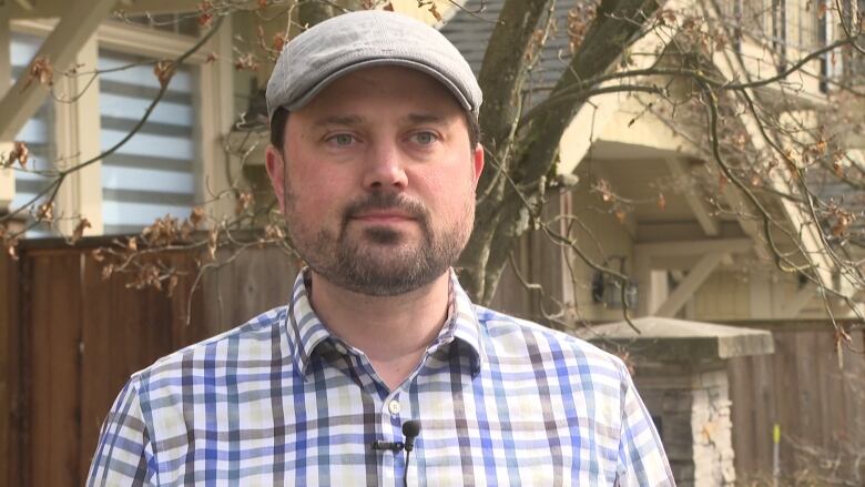 A man with a beard, checkered shirt a flat cap stands outside in a residential neighbourhood.