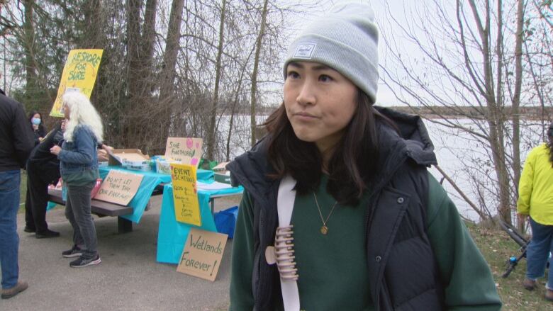 A woman with short brown hair and a grey beanie stands outside at a protest.