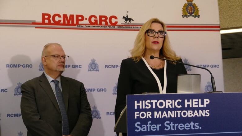 A man wearing glasses, a dark suit and dark tie stands to the left of a woman with blond hair wearing a black jacket who is standing in front of a microphone podium.