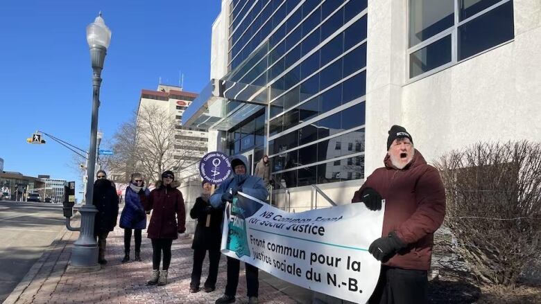 Four people stand on a sidewalk in winter coats, two people are holding a white banner that says N.B. Common Front for Social Justice in black lettering.