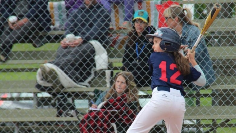 A girl wearing white pants and a blue jersey stands ready at bat as parents watch from the stands. 