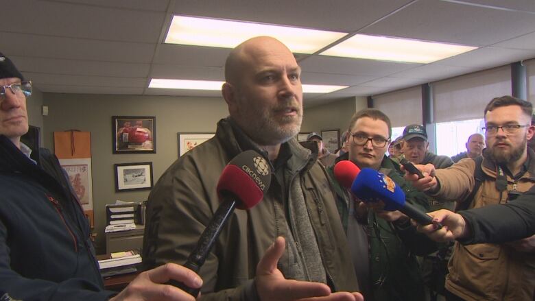 A bald man wearing a green coat stands in front of several microphones.