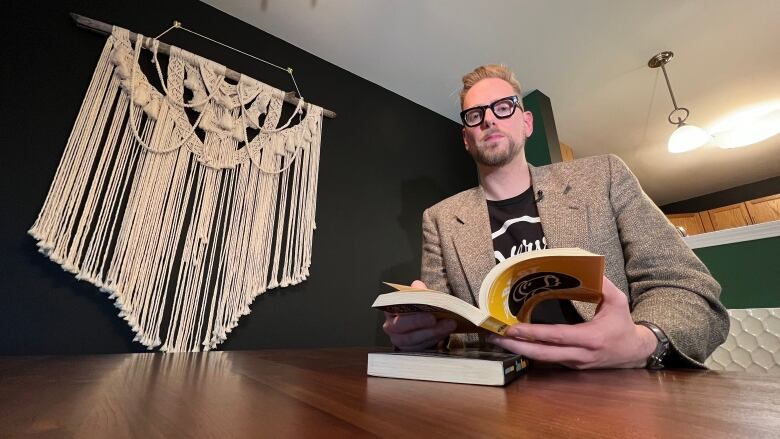 A man, Andrew Unger, poses for a photo while seated at his dining room table. He is holding an open copy of his book, The Best of the Bonnet.