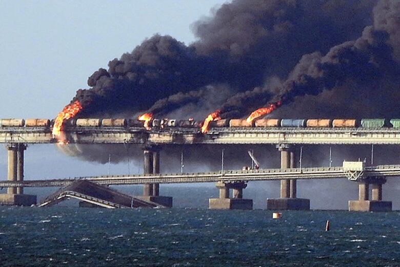 Smoke billows from a train on a bridge, which also shows several box cars in flames. 