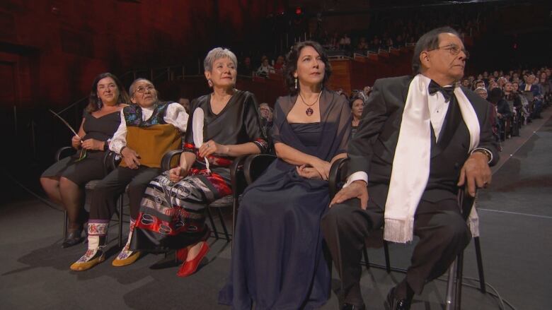 Five people in formal clothing sit in the front row of a filled auditorium. 