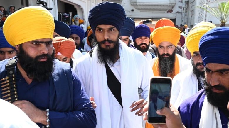 A man with a long black beard, wearing white clothing a navy blue turban, walks in a crowd of other men. 