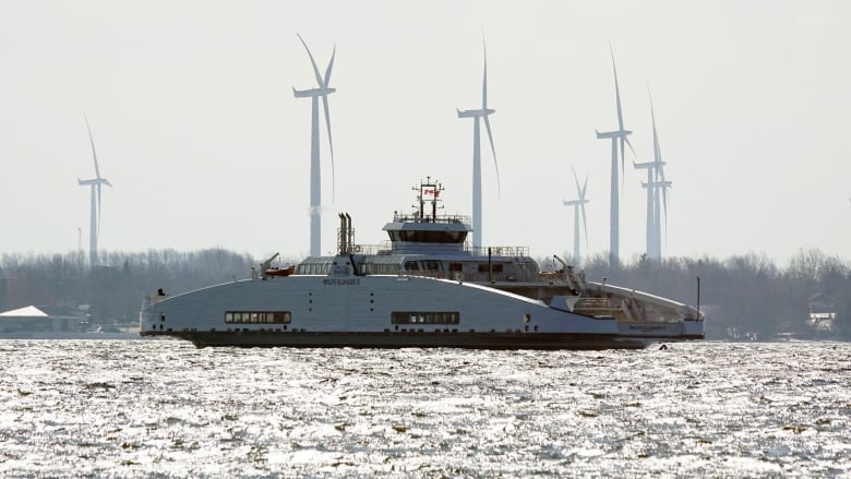 A white ferry flying a Canadian flag can be seen floating on the water. Trees and wind turbines can be seen on an island behind it.