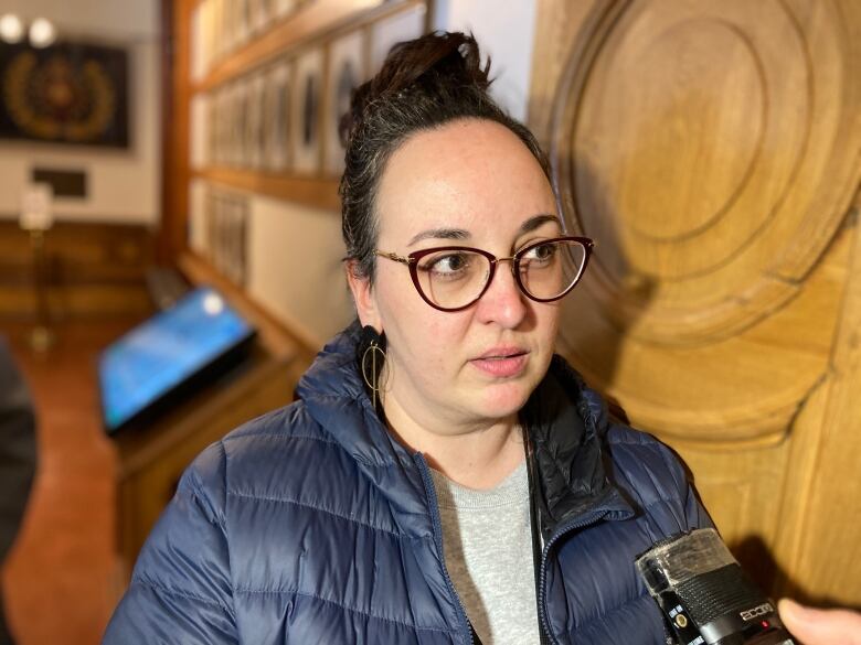 A woman with brown hair in a bun and glasses talks to a reporter's microphone.