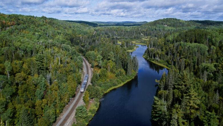 Aerial shot of a two-car Budd train snaking along a river, with endless forests and small mountains in the background.