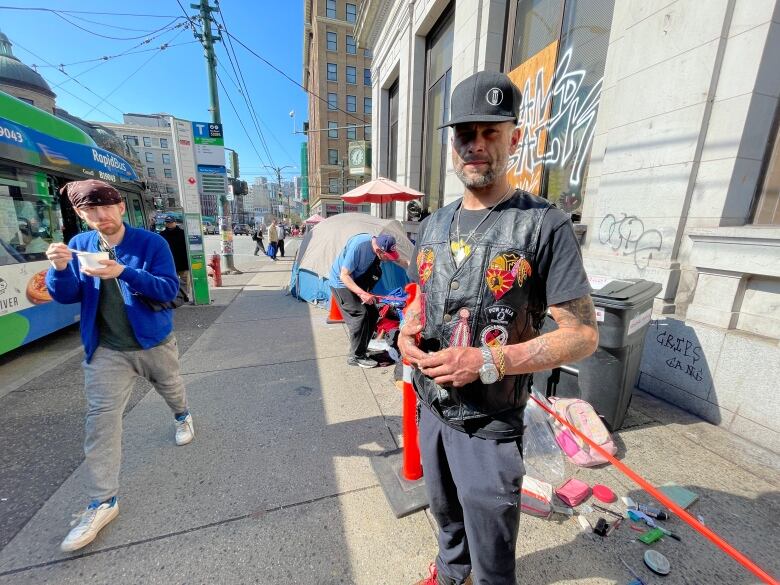 A man with a hat, black vest with a T-shirt underneath stands on a Downtown Eastside sidewalk on a sunny day.