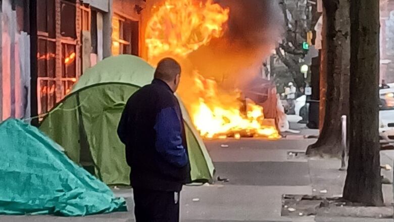 A man stands a few metres away from a tent on fire. 