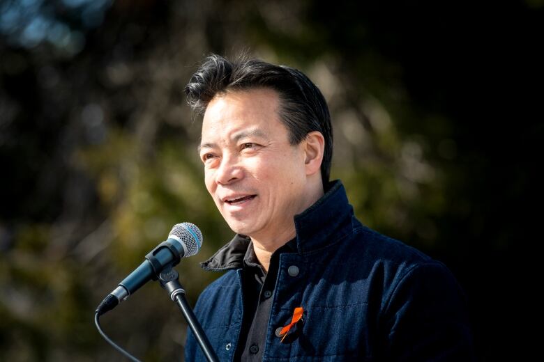 A man in blue jacket and red ribbon speaks in front of a microphone.