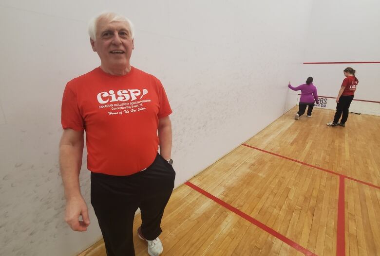 A middle-aged man stands in front of a white wall in a squash court. In the background an assistant coach guides a squash player through a warm-up. 