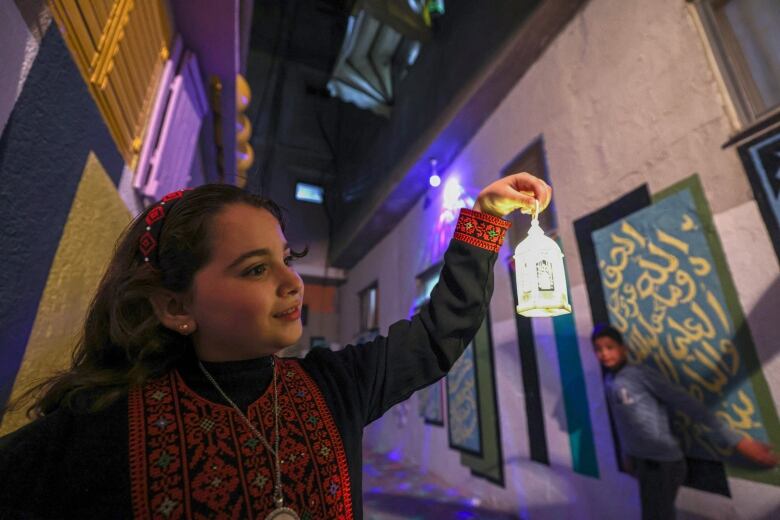 A girl holds a small traditional lantern with her left hand, with a child watching in the background standing against a wall