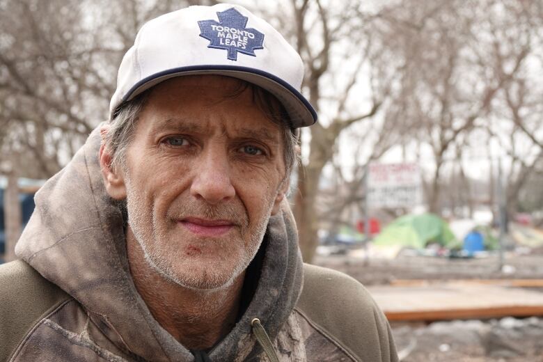 A man with a tanned, wrinkled face and a cut on his chin stares solemnly at the camera. He's wearing a camouflage hoodie and Maple Leafs ball cap.