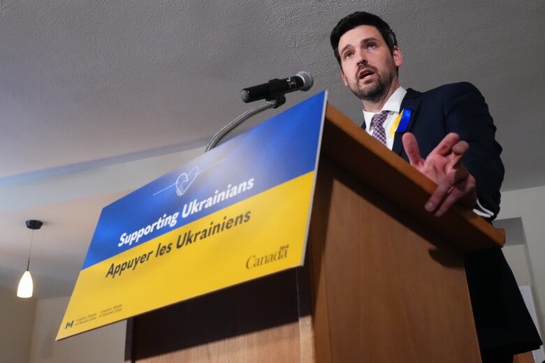 A man in a suit stands at a podium that bears the Ukrainian flag.