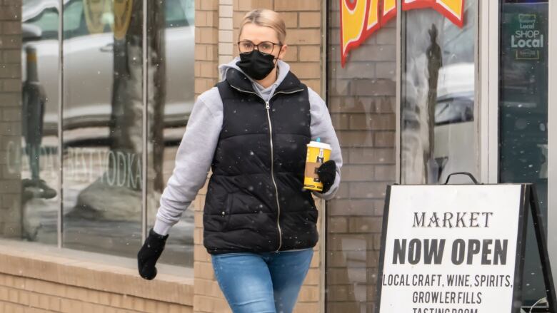 A woman wearing a black face mask walks down a city street, holding a coffee cup, as light snow falls.