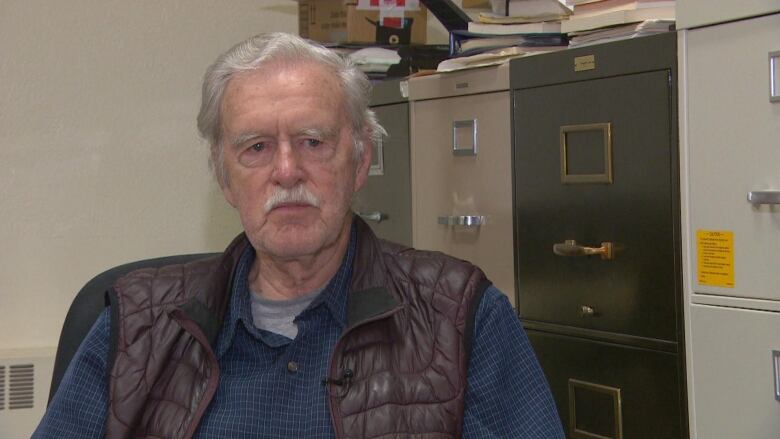 An old man with a white moustache sits in a blue shirt and brown half jacket.