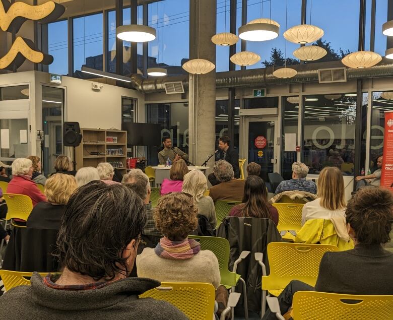 People sit in yellow and green chairs, watching two people speaking at the front of a room.