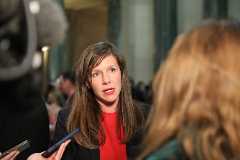 A woman with long brown hair, wearing a black blazer over a red crewneck shirt, speaks to reporters in a rotunda.