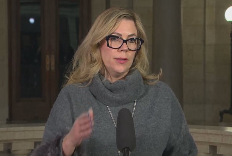 Woman with blond hair and glasses stands at a microphone in the rotunda of the legislature.