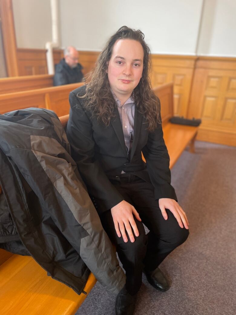 A young man with long hair wearing a dark suit. He's sitting on a bench in the viewing gallery of a courtroom.