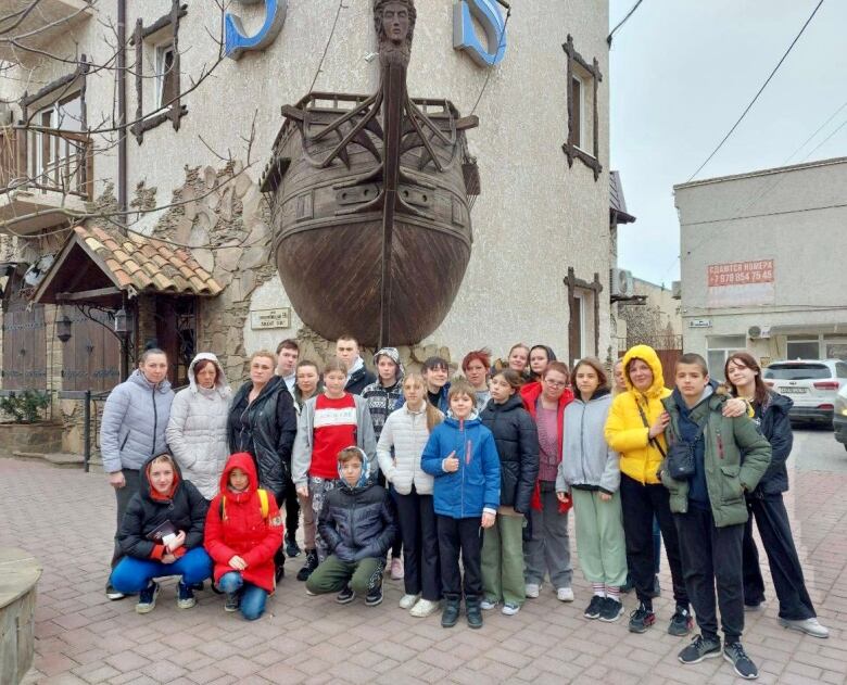 A group of adults and children wearing winter clothing pose for a photo under a historic-looking building with a ship sculpture on a brick road.