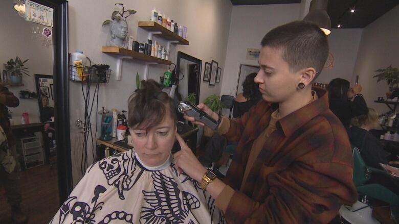 A hairdresser with close-cropped dark hear and wearing a brown plaid shirt uses a razor on a client sitting in a chair.