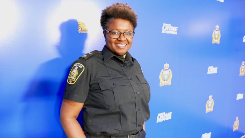 Black female police officer with black-rimmed glasses and curly hair stands in her uniform in front of a blue wall with the Winnipeg Police Service logo.