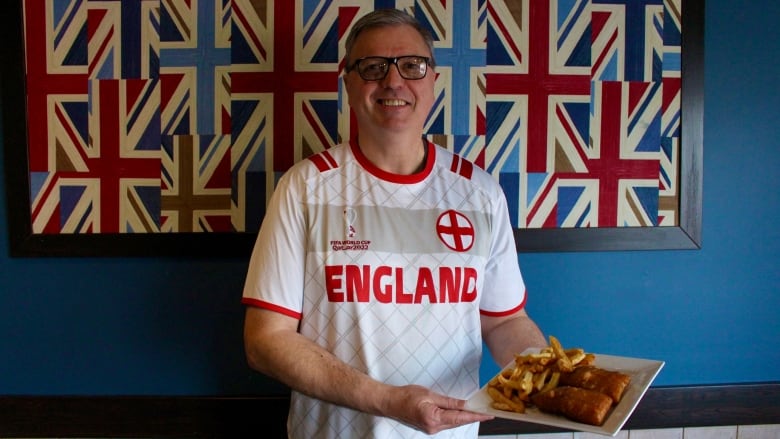 A man wearing a white and red England FIFA World Cup 2022 jersey stands in front of framed artwork featuring multiple stylized Union Jack flags.