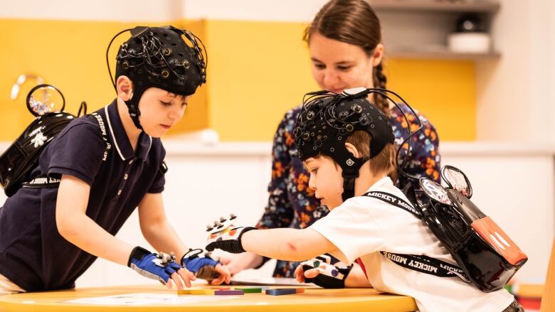 Two children sit across the table from each other, doing a puzzle-type activity. They are both wearing caps with wires coming out of them, which are used to monitor brain activity. 