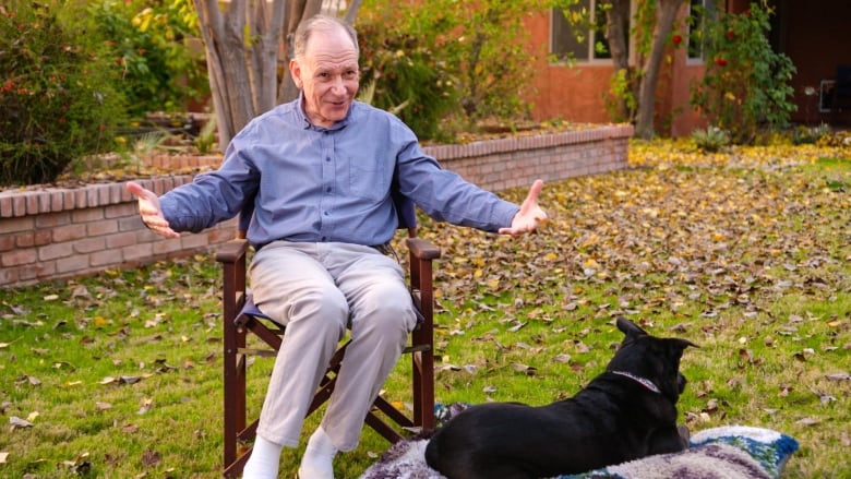 Clive Wynne, a white man wearing khakis and a blue button-down short, sits with arms outstretched in a chair next to a large black dog, who is facing away from the camera. 