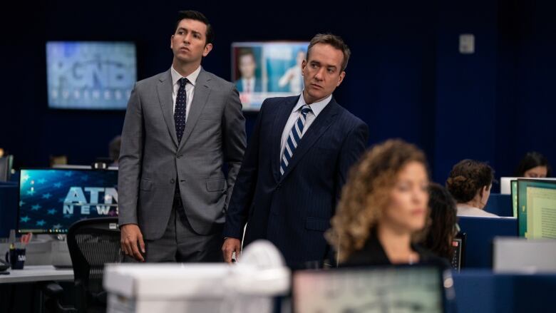 Two men, one wearing a gray suit and the other a navy suit, stand in a newsroom.