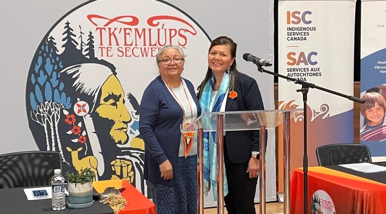 Two women smile and stand together in front of a backdrop with the Tk'emlps te Secwpemc logo.