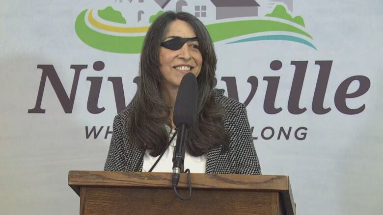 A woman with long hair and an eyepatch speaks at a podium.