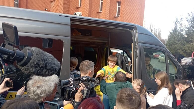 A father reaches for his smiling son as the child exits a van, amid a crow of people and news cameras.