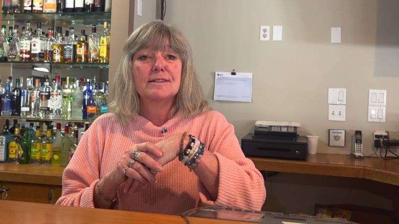 A woman speaks to the camera with alcohol bottles positioned behind her.