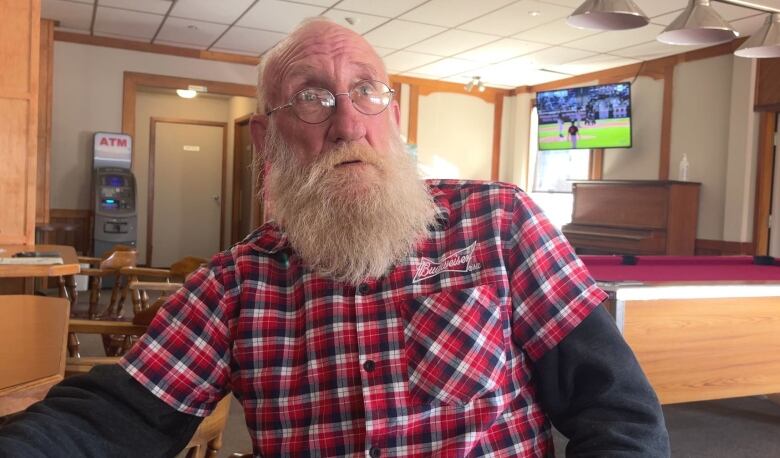 A man wearing a checkered shirt sits in a bar and talks to the camera.