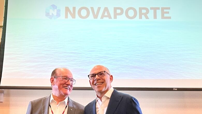 Two men with glasses smile in front of a projector screen with the company name Novaporte on it.