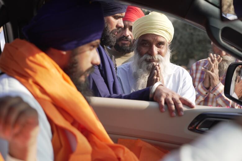 A man wearing a turban sits in a car, and through the window an elderly man smiles with his hands clasped together in front of him. 