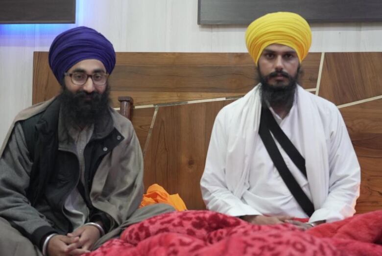 A man wearing a blue turban and glasses sits beside another man with a yellow turban on the floor. 