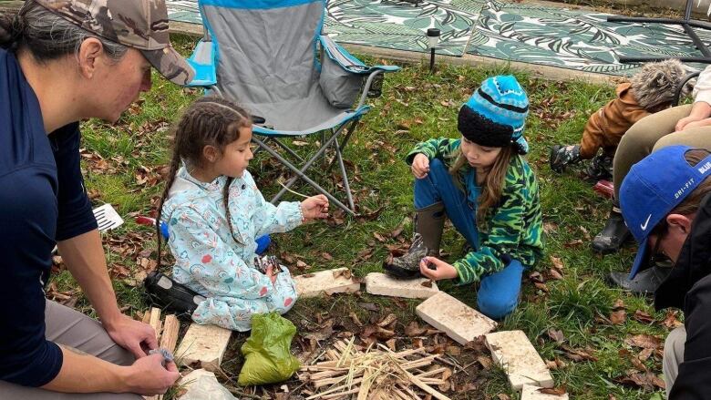 One of the parents of teaches children how to build a fire. Children sit around an unlit fire talking to each other.