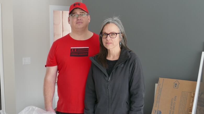 A man in a red T-shirt and baseball cap and a woman in a grey jacket stand in a room in a house that is under construction.