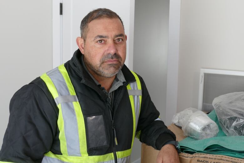 An unsmiling man in a jacket with yellow reflective safety markings stands in a room of a home.