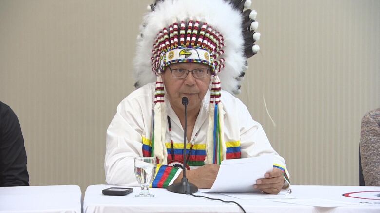 An Indigenous man wearing a headdress reads from a letter into a microphone.