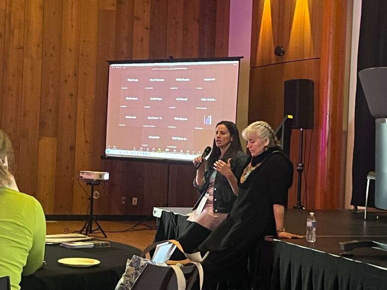 Two women lean against a stage, addressing a conference room. One holds a microphone and is speaking, gesturing with her hands.