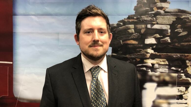 A man in a suit stands in front of a picture of an inukshuk.