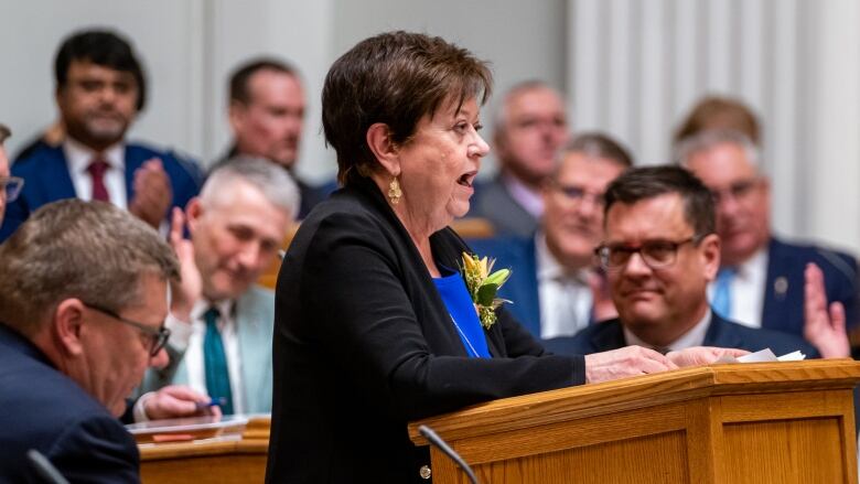 A woman with short brown hair and wearing a black suit speaks while standing at a podium surrounded by a group of men.