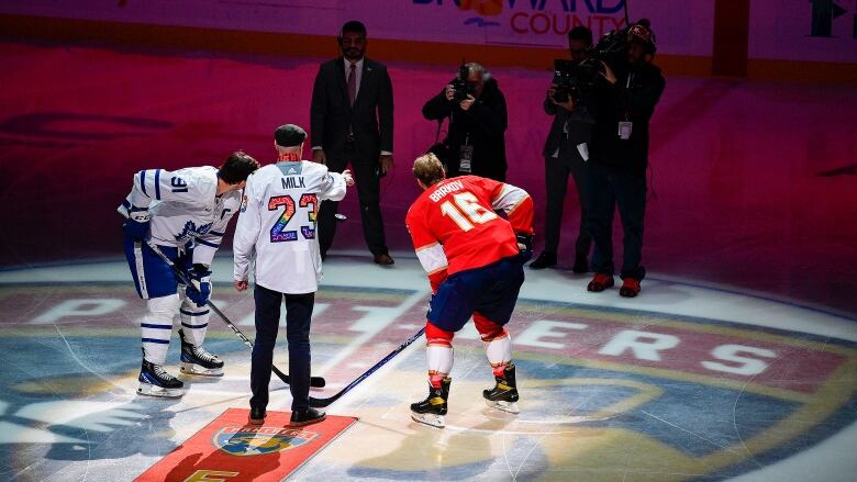 Two hockey players pose for a photograph during a faceoff, while someone drops the puck. Photographers watch in the background.